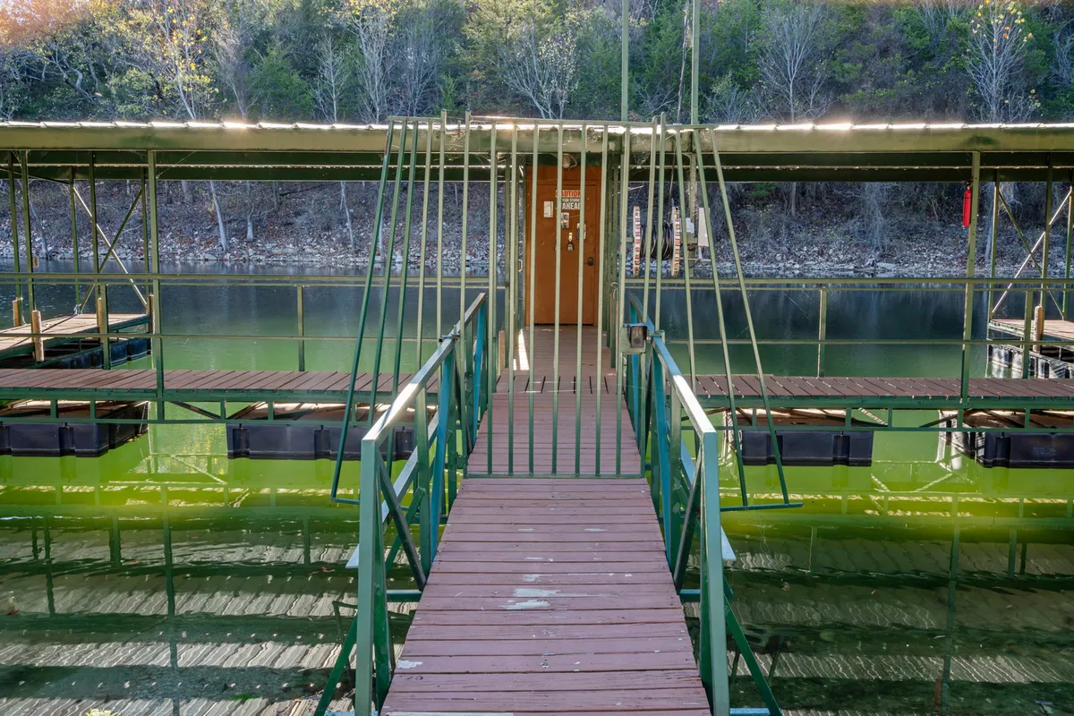 A tranquil wooden walkway with green railings leads directly to the water's edge, framed by lush trees. The calm water surface reflects the soft light, hinting at serene mornings and picturesque sunsets. The entrance door is visible, emphasizing easy access from lodging to nature.