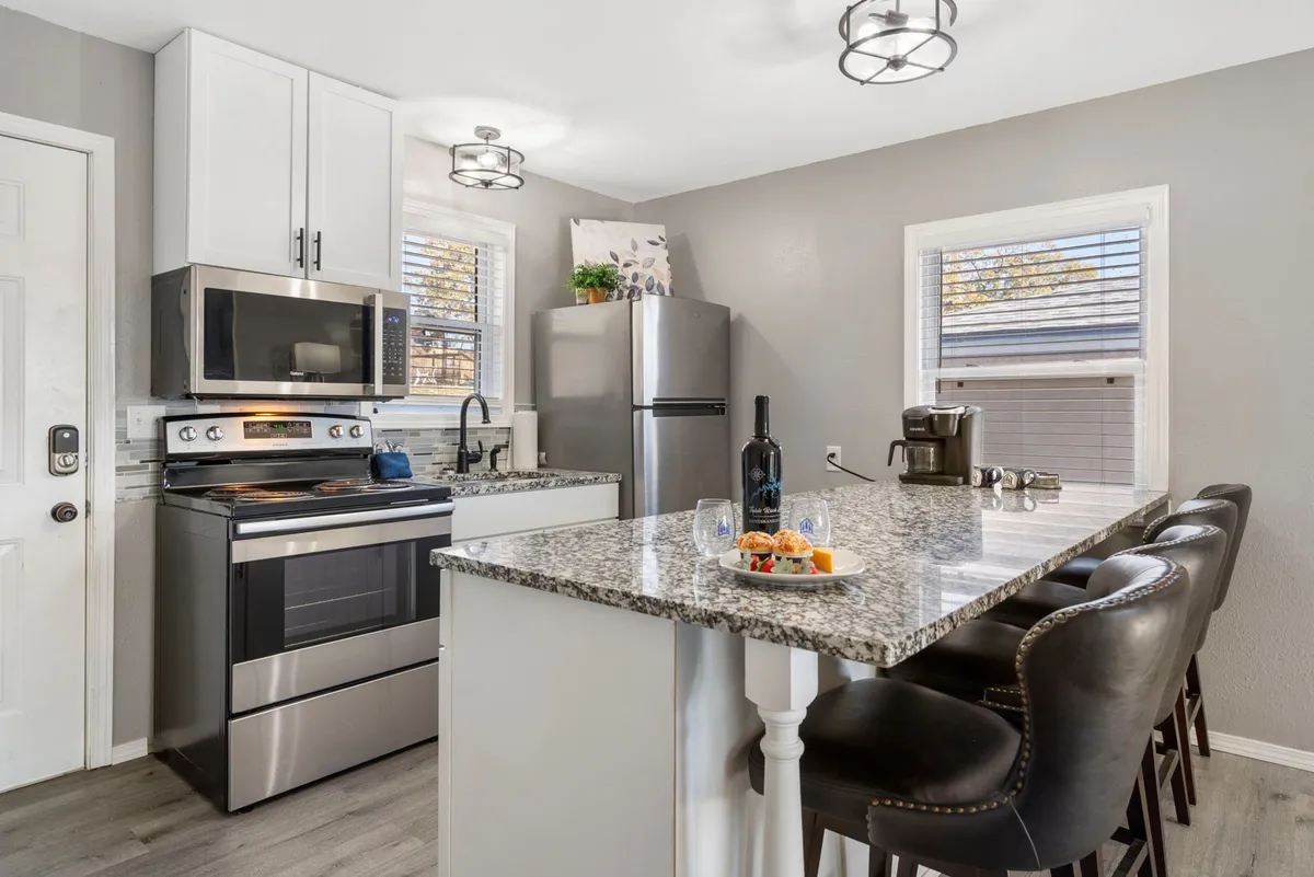 The image showcases a bright, modern kitchen featuring a spacious granite island countertop, sleek stainless steel appliances, bar stools for casual seating, and a touch of greenery from a decorative plant. The inviting setup encourages cooking, socializing, and enjoying meals together.