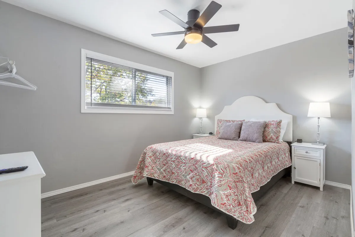 A cozy bedroom scene with patterned bedding on the bed, a ceiling fan overhead, a nightstand with a lamp, and a window with blinds. This room feels relaxed and inviting, emphasizing comfort and quiet moments.