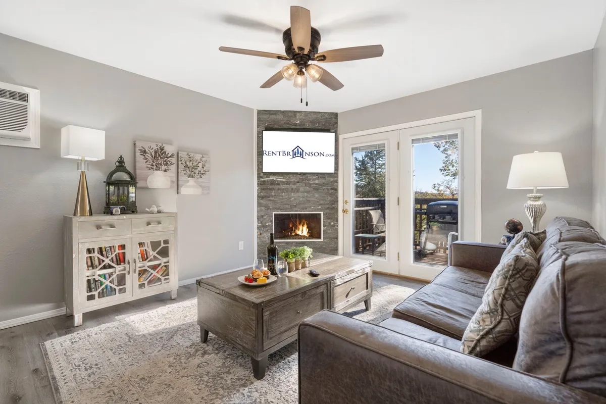 A bright, inviting living area with a plush sofa, coffee table, and a prominent fireplace. Sliding glass doors open to what appears to be a lakeside deck, and large windows flood the room with natural light. The overall layout is designed for both relaxation and entertaining, with indoor-outdoor flow.