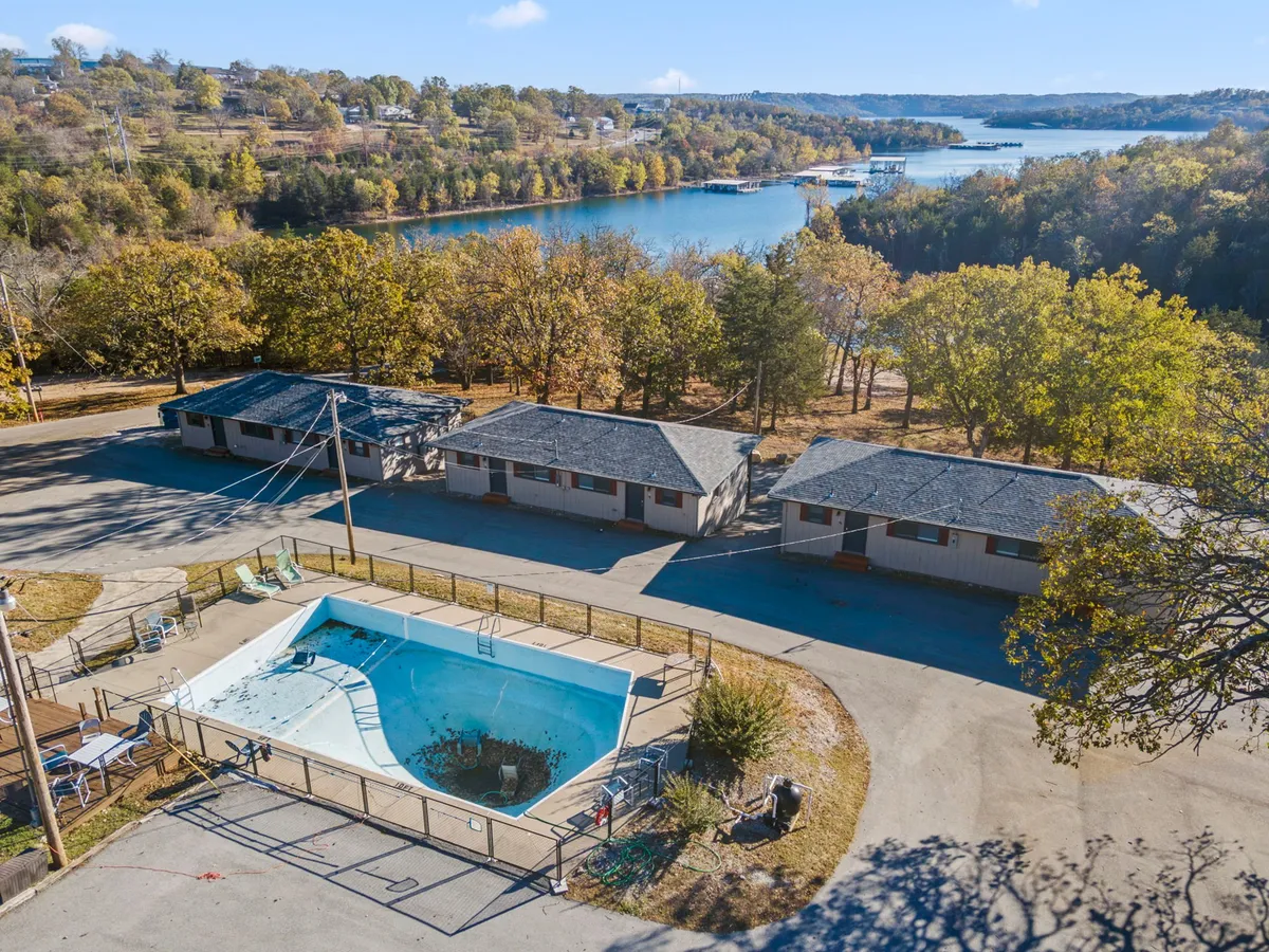 A view highlighting a swimming pool, lush trees, a clear lake in the background, and motel units nearby. The space merges poolside leisure with lakeside exploration, suggesting a wide range of outdoor activities in a pet-friendly environment.