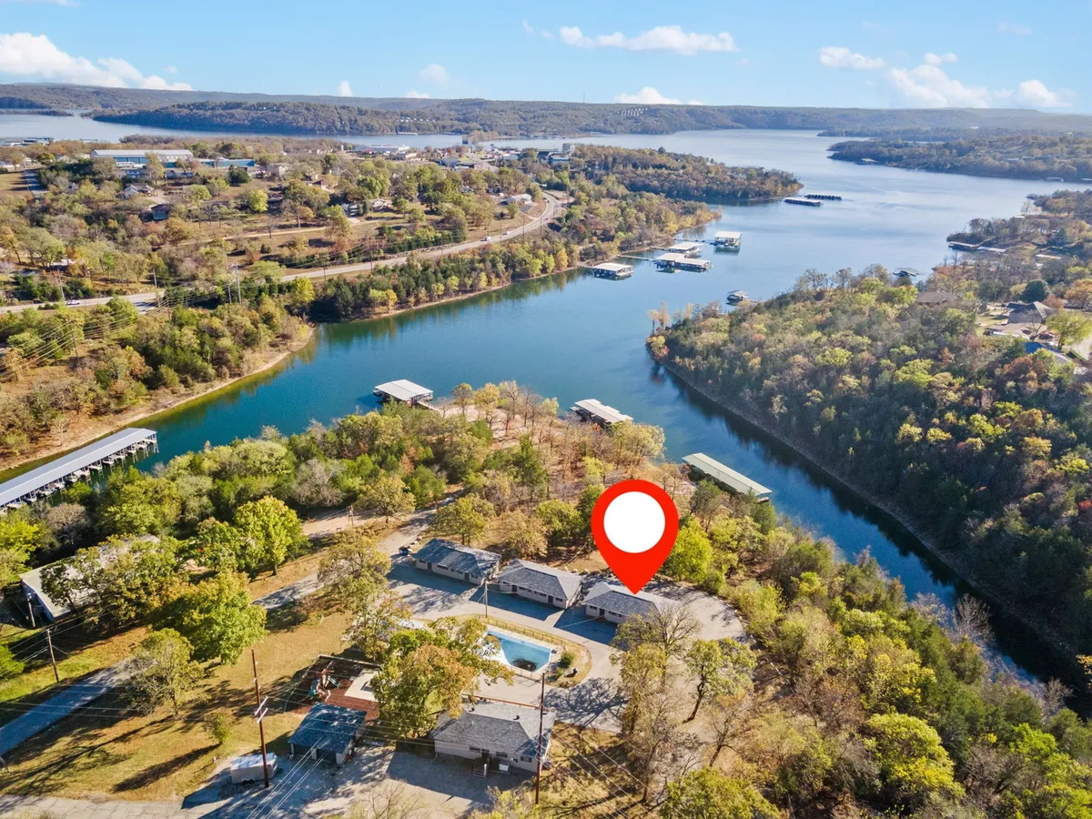 A serene lakeside setting showing the water bordered by trees, adjacent homes, and a visible pool, all under warm natural light. The atmosphere is relaxed and outdoorsy, with the landscape inviting for activities like walking, playing, or relaxing by the lake with a pet.