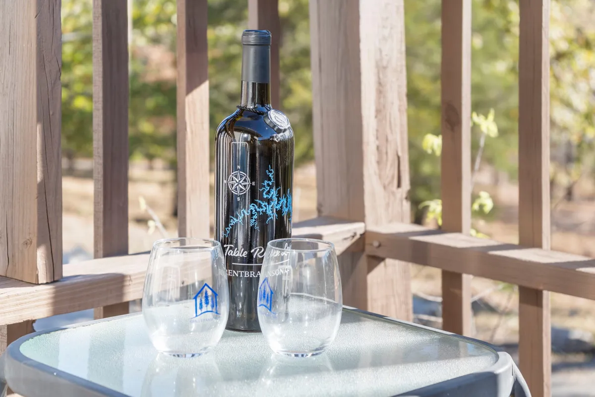 Two wine glasses and a wine bottle set on an outdoor table, positioned to overlook a scenic view—perhaps of the lake at dusk. The image suggests sharing drinks at sunset, conversation, and creating romantic or memorable moments in an outdoor setting.