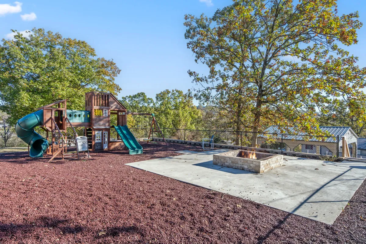 A cheerful playground structure stands adjacent to a welcoming fire pit, both set amidst leafy trees. The setup suggests lively, multi-generational gatherings, from playful adventures to cozy evenings by the fire.