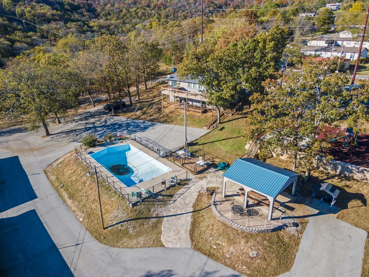 An inviting exterior pool scene with a sparkling swimming pool, a shaded gazebo, and mature trees surrounding the area. Houses are visible in the background, reinforcing the community aspect and accessibility of the amenities.