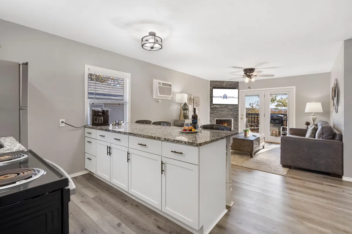 This image presents an open-concept living area where the kitchen seamlessly flows into a cozy living space. The scene features a granite kitchen island with seating, a fireplace, comfortable living room seating, and large windows that suggest abundant natural light and a calming view beyond. The arrangement encourages group gatherings, cooking, and relaxation.