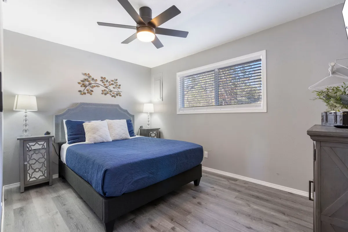 This image showcases a bedroom with a blue bedspread, decorative wall art above the bed, nightstands with lamps on either side, and a large window letting in natural light. The setup exudes tranquility and comfort, emphasizing soft bedding and restful décor.